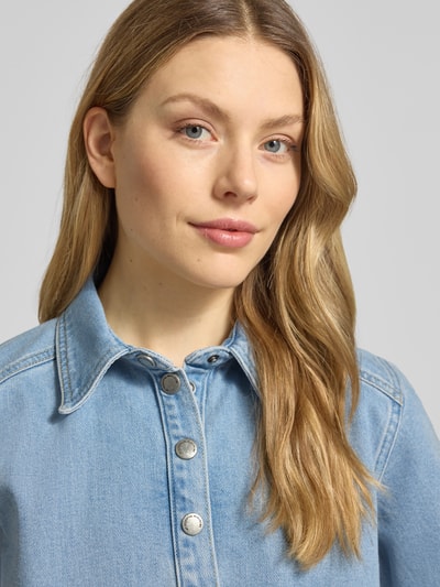 Modelka nosi bluzkę jeansową z mieszanki bawełny z dodatkiem streczu, pudełkowy krój, listwa z zatrzaskami, model 'Abina' od My Essential Wardrobe, kołnierzyk typu kent, jasnoniebieski materiał, wyraźna faktura denimu, damska moda, zbliżenie górnej części ciała.