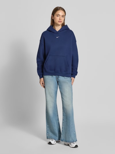 Frauenmodel trägt Oversized Hoodie aus Baumwoll-Mix von Nike in Dunkelblau, kombiniert mit hellblauen Jeans, Ganzkörperansicht von vorne. Damen Hoodie von Nike.