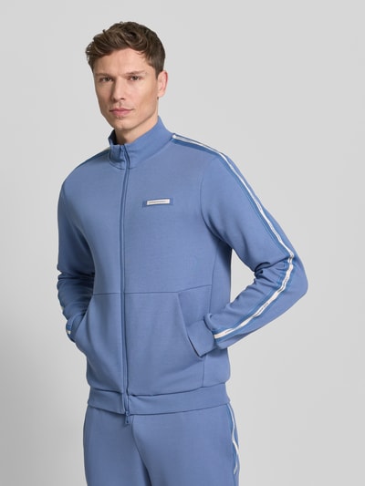 Männermodel trägt Sweatjacke aus Baumwoll-Mix von EA7 Emporio Armani in Blau mit Stehkragen, Galonstreifen und Känguru-Tasche, Nahaufnahme Oberkörper.