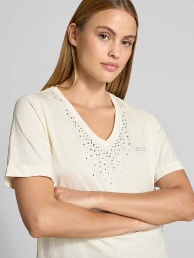 Frauenmodel trägt T-Shirt mit Strasssteinbesatz und V-Ausschnitt von LIU JO SPORT in Creme, Baumwolle, gerader Schnitt, Nahaufnahme Oberkörper.
