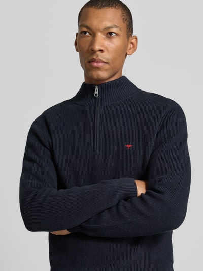 Męski model nosi Regular Fit Strickpullover aus reiner Baumwolle mit Troyer-Kragen od Fynch-Hatton, granatowy ze strukturalnym splotem, logo z przodu, długie rękawy, żebrowane wykończenia, widok na górną część ciała z przodu, kontekst: sweter męski, czysta bawełna, krój regular fit, stójka z zamkiem, długość tyłu w rozmiarze M: 65 cm, długość rękawa w rozmiarze M: 75 cm.