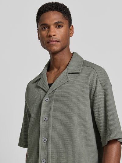 Męski model nosi Relaxed Fit Freizeithemd mit Strukturmuster od Drykorn z bawełny, z kołnierzem z połami, z krótkim rękawem, fakturowany wzór, listwa guzikowa, kolor khaki, długość tyłu w rozmiarze M: 69 cm, szerokość w rozmiarze M: 60 cm, długość rękawów w rozmiarze M: 26 cm, fakturowana powierzchnia, widok na górną część ciała z przodu.
