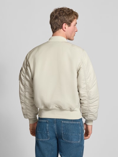 Männermodel trägt Regular Fit Bomberjacke mit Stehkragen Modell 'Heritage' von Alpha Industries in Beige mit elastische Abschlüsse, Rückenlänge 63 cm, Schulterbreite 50 cm, Brustweite 57 cm, Pattentaschen, Reißverschluss, glatte Struktur, Ganzkörperansicht Rückansicht.