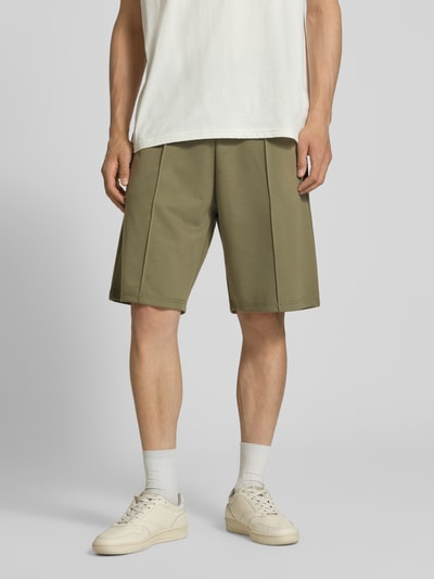 Männermodel trägt Shorts mit elastischem Bund und Ziernähten von REVIEW, Loose-Fit Material-Mix mit Stretch-Anteil, Bundweite 42 cm, Innenbeinlänge 26 cm, olivegrün, seitliche Eingrifftaschen, angedeutete Gesäßleistentaschen, Ganzkörperansicht von vorne.