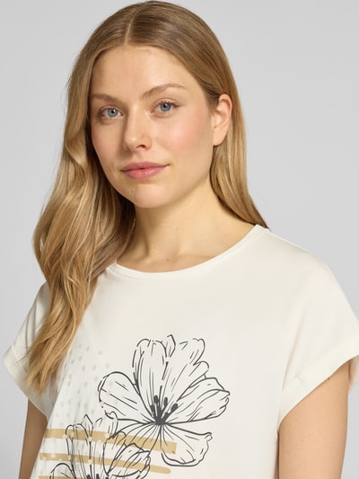 Modelka kobieca nosi T-Shirt mit fixiertem Ärmelaufschlag Modell 'RINA' od Soyaconcept, kremowe z motywem kwiatowym, bawełniany miks z domieszką stretchu, krój prosty, okrągły dekolt, detaliczne zbliżenie na górę ciała.