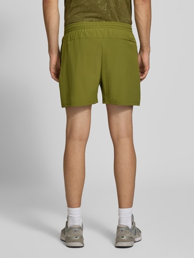 Männermodel trägt Shorts mit elastischem Bund und Logo von New Balance in Olivgrün, lockere Passform, Rückansicht, Ganzkörperansicht von hinten.
