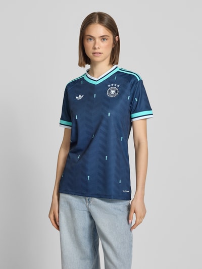 Modelka kobieca nosi wielokolorowe DFB Auswärtstrikot 2026 od adidas Sportswear z krótkim rękawem, krój regular fit, V-neck, aplikacja logo DFB i label stitching, faktura materiału z geometrycznymi wzorami, widok całej sylwetki z przodu.