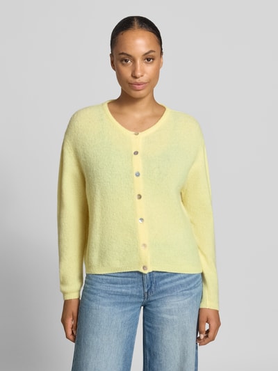 Frauenmodel trägt Relaxed Fit Strickjacke aus Woll-Mix mit Leinen-Anteil von American Vintage in zartem Gelb, gerippte Bündchen, durchgehende Knopfleiste, Langarm, Ganzkörperansicht von vorne.