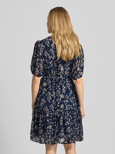 Kobiece modelka nosi Knielanges Kleid mit Kordelzug an der Taille od Apricot, granatowy kolor, wzór liści, długość do kolan, desenie, rękaw o dł. 1/2, taliowany fason, zakończenia rękawów elastyczne, dekolt w serek, dół obszyty falbaną, długość tyłu w rozmiarze S: 93 cm, szerokość w rozmiarze S: 43 cm, długość rękawów w rozmiarze S: 30 cm, widok całej sylwetki z tyłu.