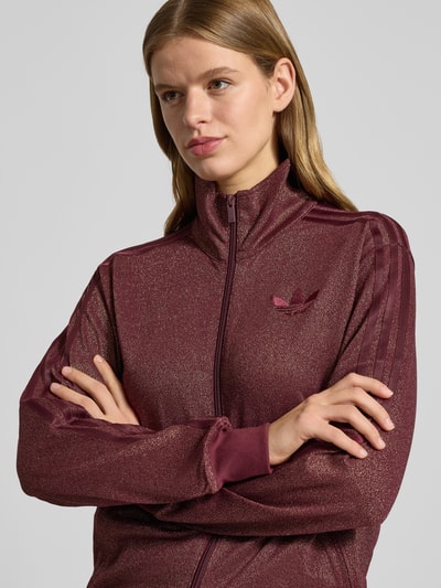 Kobieta modelka nosi bluzę rozpinaną z stójką od adidas Originals, mieszanka bawełny z metalem, luźny krój, zamek błyskawiczny, wyhaftowane logo, prążkowane wykończenia, kieszenie zapinane na zamek, bordowy kolor, faktura z połyskiem, zbliżenie na górną część ciała.