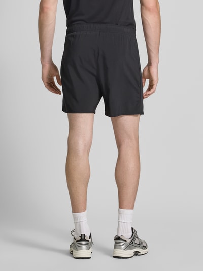 Męski model nosi Regular Fit Shorts mit integriertem Slip Modell 'ICON' od ASICS TIGER, czarny kolor, szerokość w rozmiarze M: 37 cm, długość wewnętrzna nogawki w rozmiarze M: 18 cm, nadruk z logo, elastyczny pas, krój regular fit, całkowity widok z tyłu.