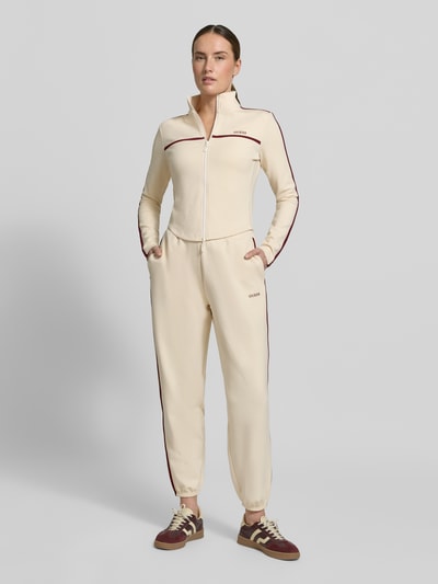 Modelka kobieca nosi Jogpants mit Logo Modell 'JASMINE' od Guess Activewear, beżowy materiał z Lyocellem, krój regular fit, kieszenie, widok całej sylwetki z przodu.