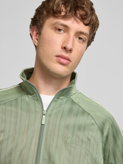Männermodel trägt Regular Fit Sweatjacke mit opstaande kraag van Karl Kani in lichtgroen met ritssluiting, elastisch; nahaufnahme bovenlichaam. Ruglengte maat M: 70 cm, mouwlengte maat M: 67 cm.