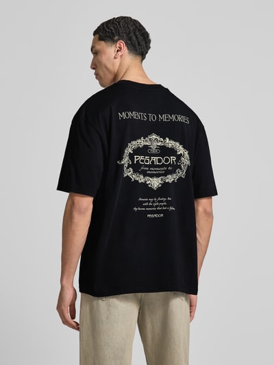 Männermodel trägt Oversized T-Shirt mit rückseitigem Motiv-Print Modell 'Reine' von Pegador in Schwarz, Reine Baumwolle, Rundhalsausschnitt, Oversized-Fit, Rückenlänge 70 cm bei Größe M, Label-Print, Ganzkörperansicht Rückansicht.