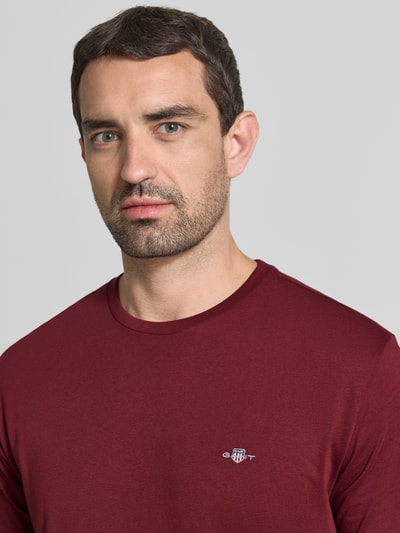 Mannenmodel draagt Regular fit t-shirt uit puur katoen van GANT in bordeauxrood, logo op de borst, close-up bovenlichaam.
