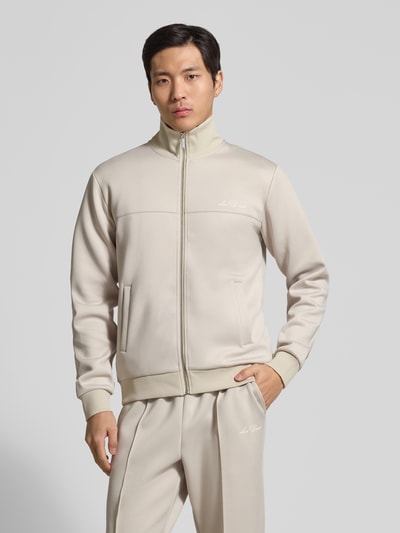 Męski model nosi Sweatjacke mit Logo-Print Modell 'Ballier' von Les Deux, beżowy materiał z domieszką stretchu, prosty krój, stójka, zamek, boczne kieszenie, ściągacze, długość tyłu w rozm. M: 67 cm, długość rękawa: 68 cm, widok całej sylwetki z przodu.