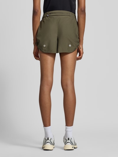 Frauenmodel trägt ASICS TIGER Shorts mit elastischem Bund Modell 'NAGINO' in Olivgrün, reflektierende Details, Rückansicht Ganzkörperansicht.