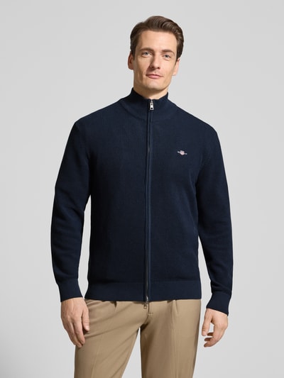 Männermodel trägt Cardigan mit Zweiwege-Reißverschluss und Label-Stitching von Gant aus reiner Baumwolle, Slim-Fit, Stehkragen, dunkelblau, beige Hose, Ganzkörperansicht von vorne.