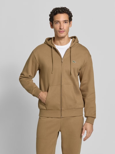 Männermodel trägt Regular Fit Sweatjacke aus Baumwoll-Mix von Lacoste in Beige mit Reißverschluss und Kängurutaschen, Ganzkörperansicht von vorne. Herren Sweatjacke von Lacoste.