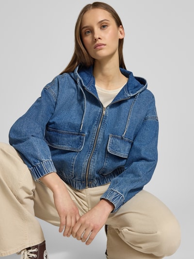 Modelka kobieca nosi Regular Fit Jeansjacke aus reiner Baumwolle Modell 'KENZIE' od Only, denim, krój regular fit, długi rękaw, elastyczne mankiety i dół, patkowe kieszenie, stójka z kapturem i troczkiem, zapięcie na zamek, materiał bawełna; kolory niebieski i beżowy; pełny widok z przodu.