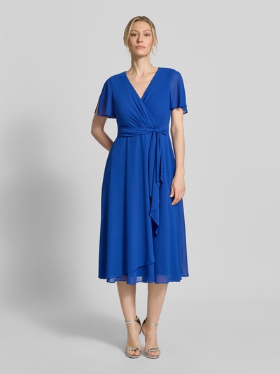 Kobiece model prezentuje Knielanges Cocktailkleid mit Bindegürtel von Christian Berg Cocktail z szyfonu, taliowany, długość do kolan, kopertowy styl, wiązany pasek, długość tyłu w rozmiarze 36: 111 cm, szerokość w rozmiarze 36: 38 cm, długość rękawów w rozmiarze 36: 22 cm; intensywny niebieski kolor, gładka tekstura, widok całej sylwetki z przodu.