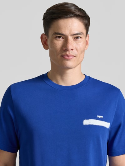 Männermodel trägt T-Shirt mit rückseitigem Motiv-Print Modell 'Bobby' von Wood Wood in Blau, Baumwolle, Logo-Print, Nahaufnahme Oberkörper.