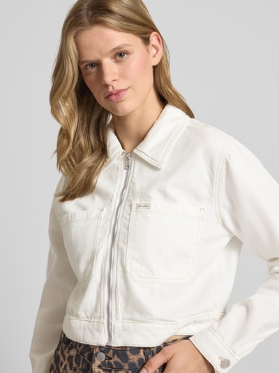 Frauenmodel trägt Cropped Jeansjacke mit Brusttaschen und Label-Patch von Guess Jeans aus Baumwoll-Lyocell-Anteil, Straight Fit, Reißverschluss, weiße Farbe mit sichtbarer Struktur, Nahaufnahme Oberkörper.