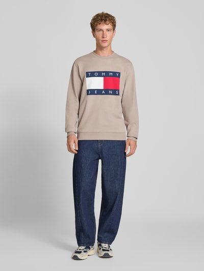 Męski model nosi Regular Fit Sweatshirt aus reiner Baumwolle von Tommy Jeans, beżowa bawełna, granatowe dżinsy, Ganzkörperansicht z przodu