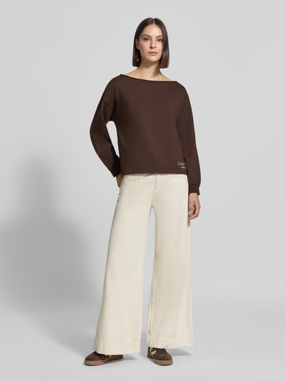 Frauenmodel trägt Relaxed Fit Pullover aus Baumwoll-Mix von Smith and Soul in Dunkelbraun mit Statement-Stitching, helle Wide-Leg-Hose, Ganzkörperansicht von vorne.