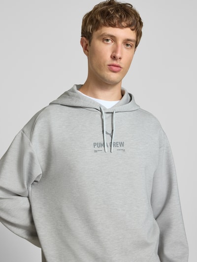 Männermodel trägt Hoodie mit Logo von PUMA PERFORMANCE in Hellgrau, Relaxed Fit, Kapuze, Material-Mix, Nahaufnahme Oberkörper.