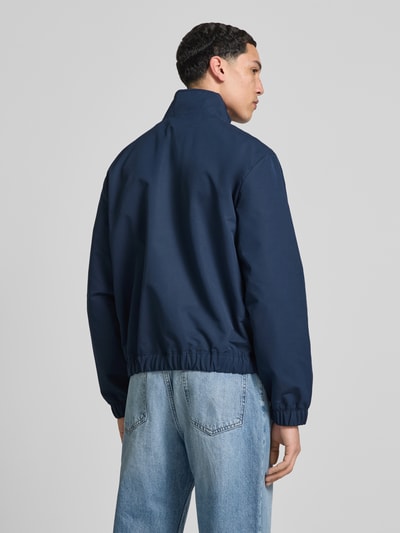 Männermodel trägt Regular Fit Blouson mit stójką od Tommy Jeans, efekt: jednolity kolor, długi rękaw, wpuszczane kieszenie, naszywka z logo, krój regular fit, elastyczne wykończenia, kieszeń wewnętrzna, zamek błyskawiczny z przodu, widok całej sylwetki od tyłu.