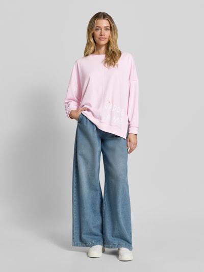 Frauenmodel trägt Oversized Sweatshirt mit Statement-Print und Rollsaum von Smith and Soul aus Baumwoll-Mix mit Modal-Anteil in zartem Rosa, kombiniert mit weiten Jeans, Rundhalsausschnitt, deutlich sichtbarem Statement-Print und markantem Rollsaum, Oversized-Fit und überschnittene Schultern, Ganzkörperansicht von vorne.