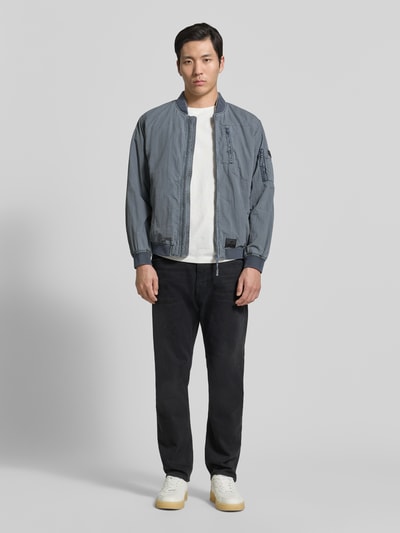Männermodel trägt Blouson mit Label-Patches Modell 'IBE' von khujo in Blau, Gerader-Schnitt, Stehkragen, Reißverschluss, seitliche Eingrifftaschen, Brusttasche mit Reißverschluss, Reißverschlusstasche am Ärmel, glatte Oberfläche, Ganzkörperansicht von vorne.