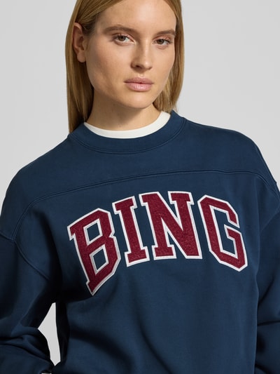 Frauenmodel trägt Oversized Sweatshirt mit Logo-Detail von Anine Bing aus reiner Baumwolle, dunkelblau mit Rundhalsausschnitt, Nahaufnahme Oberkörper.