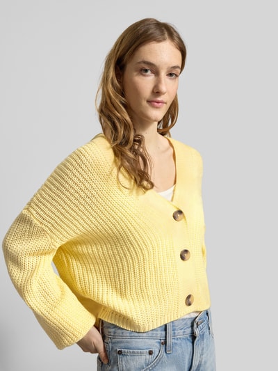 Frauenmodel trägt Regular Fit Strickjacke mit einreihiger Knopfleiste Modell 'CAROL NICE' von Only, unifarben gelb, Strickoptik, cropped, V-Ausschnitt, langarm, gerippte Abschlüsse, Ganzkörperansicht von vorne.