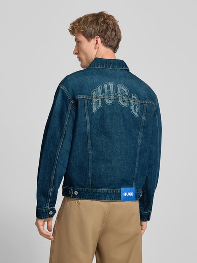 Mężczyznę model nosi kurtka jeansowa Regular Fit Jeansjacke aus reiner Baumwolle Modell 'Dex' von HUGO, niebieski dżins, beżowe spodnie, Ganzkörperansicht Rückansicht, kurtka HUGO Blue.