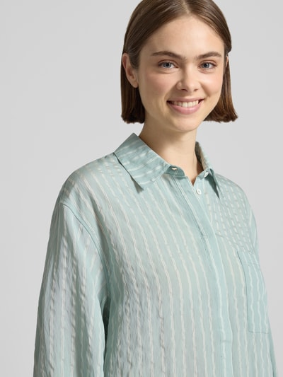 Frauenmodel trägt Bluse aus Viskose-Mix mit Brusttasche Modell 'Selina' von OH APRIL, Hemdblusenkragen, verdeckte Knopfleiste, Rüschenärmel, Rüschensaum, Hellgrün mit weißen Streifen, Struktur und Glanz, Ganzkörperansicht von vorne, Rückenlänge bei Größe S: 73 cm, Ärmellänge bei Größe S: 61 cm.
