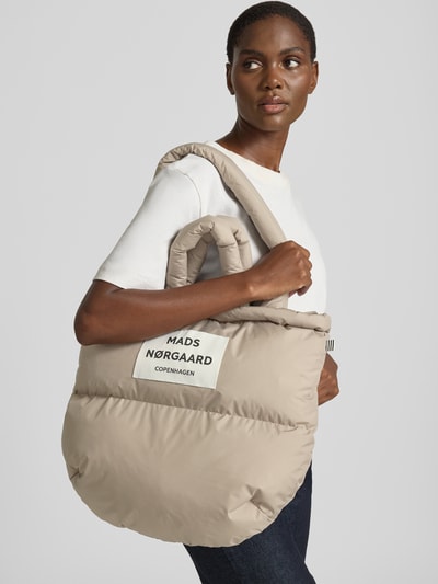 Frauenmodel trägt Handtasche mit Label-Detail und Steppnähten von MADS NØRGAARD in Beige mit Tragehenkel und Schulterriemen, Nahaufnahme Oberkörper.