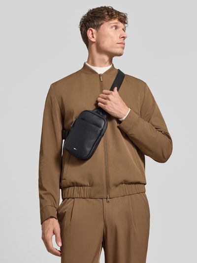 Männermodel trägt Umhängetasche mit Label-Detail Modell 'RICO Crossbody Mini' von bugatti in Schwarz, Struktur glatt, Ganzkörperansicht von vorne. Szerokość w rozmiarze 1: 14 cm, długość w rozmiarze 1: 20 cm, głębokość w rozmiarze 1: 4 cm, pasek na ramię o długości 85 cm.