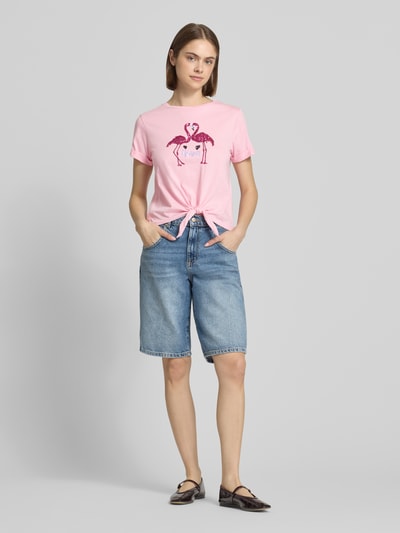 Frauenmodel trägt Regular Fit T-Shirt aus reiner Baumwolle Modell 'DORA LIFE' von Only, rosa, unifarben, Pailletten, Knotendetail, Rundhals, kurzarm, Kombi mit Jeansshorts, Ganzkörperansicht von vorne.