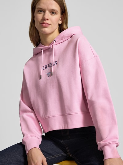 Modelka nosi Relaxed Fit Hoodie mit Logo-Stitching Modell 'Baker' od Guess Jeans, czysta bawełna, różowy, kaptur ze sznurkiem, obniżone ramiona, elastyczne wykończenia, widok na górną część sylwetki z przodu