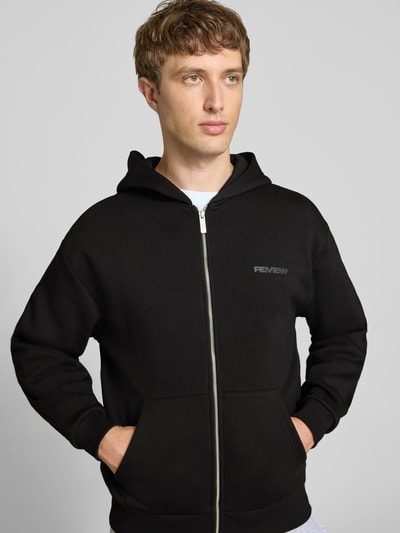 Männermodel trägt Relaxed Fit Sweatjacke aus Baumwoll-Mix mit Label-Detail von REVIEW, schwarz, Kapuze, Reißverschluss, Känguru-Tasche, elastische Abschlüsse, Ganzkörperansicht von vorne.