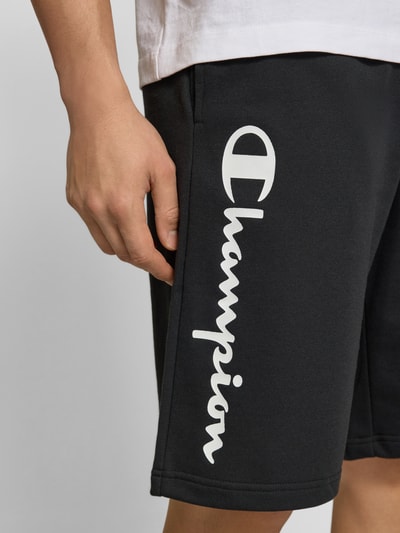 Männermodel trägt Shorts mit Logo-Print und -Stitching von CHAMPION, schwarz, Baumwoll-Mix, lockerer Schnitt, Nahaufnahme Oberschenkel.