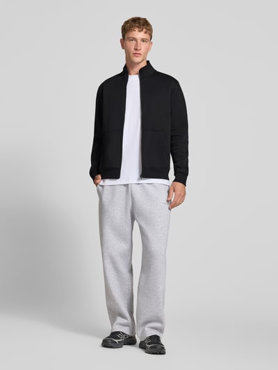 Männermodel trägt Regular Fit Sweatjacke mit Baumwoll-Anteil Modell 'Bradley' von JACK & JONES in Schwarz, Ganzkörperansicht von vorne. Material-Mix, Stehkragen, Reißverschluss, geteilte Känguru-Tasche, Rückenlänge 68 cm, Ärmellänge 66 cm.