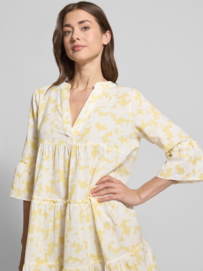 Frauenmodel trägt Ausgestelltes Blusenkleid aus reiner Baumwolle Modell 'DICTHE' von Vero Moda mit gelbem Allover-Muster, Henley-Ausschnitt und Trompetenärmeln, Mini-Länge, lockere Passform, Nahaufnahme Oberkörper.