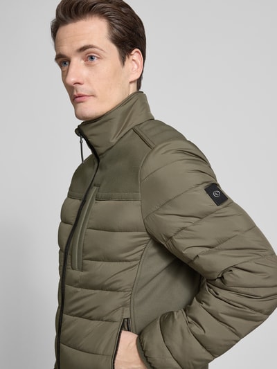 Męski model nosi Regular Fit Steppjacke mit seitlichen Stretcheinsätzen von Tom Tailor, kolor oliwkowy, naszywka z logo, stójka, długi rękaw, kieszenie na zamek, detal na plecach, materiał hydrofobowy, krój regular fit, efekt: jednolity kolor, zamek błyskawiczny, widok na górną część ciała z przodu.