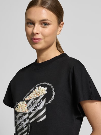 Kobiece modelka nosi Regular Fit T-Shirt mit Ziersteinbesatz Modell 'Popcorn' od Guess, bawełna, motyw nadruk, dekolt okrągły, krój regular fit, długość tyłu w rozm. S 56 cm, czarne kolory, nadruk z połyskiem, Nahaufnahme górnej części ciała.
