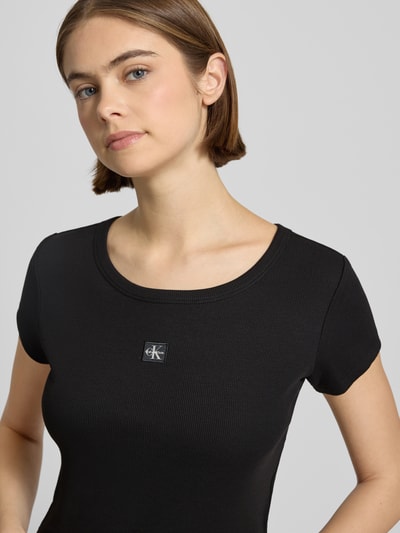 Kobiece model prezentuje Cropped T-Shirt mit Label-Detail od Calvin Klein Jeans z mieszanki bawełny i elastanu, okrągły dekolt, krótki krój, detal z logo, długość tyłu 43 cm w rozmiarze S, kolor czarny, tekstura prążkowana, zbliżenie na górną część sylwetki, widok z przodu