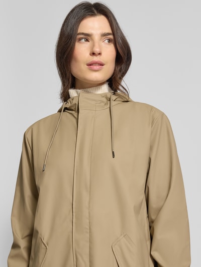 Frauenmodel trägt Regular Fit Parker mit Kapuze Modell 'Fishtail' von Rains in Beige mit Pattentaschen, elastischen Ärmelabschlüssen, Nahaufnahme Oberkörper. Rückenlänge 104 cm, Schulterbreite 45 cm, Brustweite 55 cm, Ärmellänge 65 cm bei Größe S.