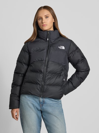Modelka nosi czarną kurtkę pikowaną z wyhaftowanym logo, zamkiem błyskawicznym i kieszeniami zapinanymi na zamek błyskawiczny, Model 'SAIKURU' od The North Face, krój regular fit, stójka, widok całej sylwetki z przodu, długość tyłu w rozmiarze S: 59 cm, szerokość ramion w rozmiarze S: 43 cm, szerokość w klatce piersiowej w rozmiarze S: 53 cm, długość rękawów w rozmiarze S: 62 cm.